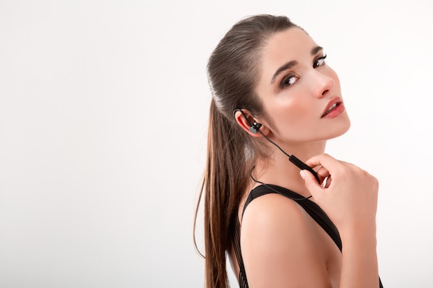 Attractive brunette woman in jogging black top listening to music on earphones posing isolated on white wall ponytail hairstyle