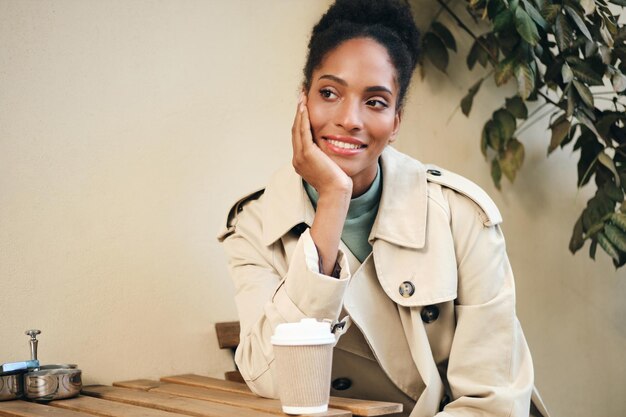 Attractive casual African American girl in stylish trench coat happily looking away with coffee to go in cafe on street