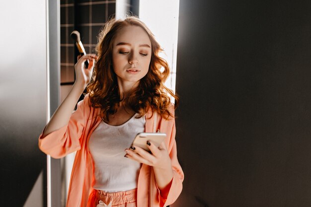 Attractive caucasian girl in pajama posing with phone at home. Indoor photo of chilling female model with wavy hair holding makeup brush.