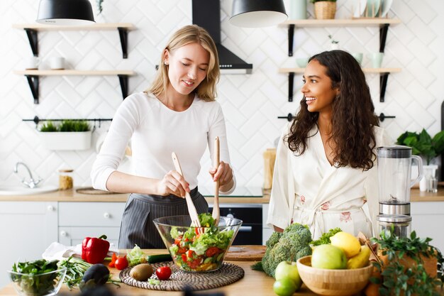 Attractive caucasian woman is cooking healthy salad and beautiful mulatto woman is looking  on her dressed in silky nightgown on modern designed kitchen