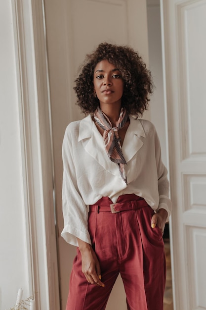 Free Photo attractive charming brunette curly woman in burgundy pants and trendy white blouse looks into camera and poses near white wooden doors