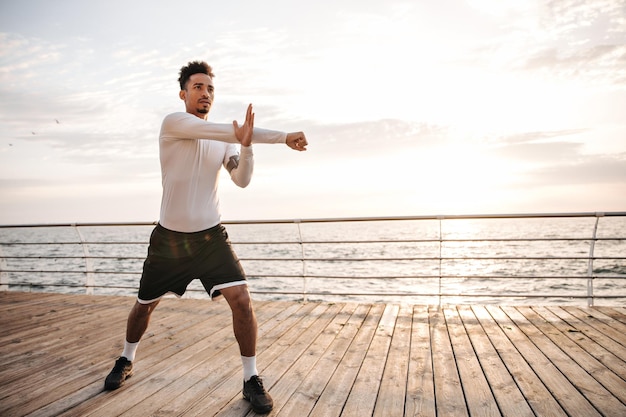 Attractive charming darkskinned sportsman in black shorts and white longsleeved tshirt does ark exercises stretches and works out near sea