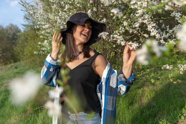 Attractive cheerful girl in a hat among the flowering trees in the spring, in a casual style