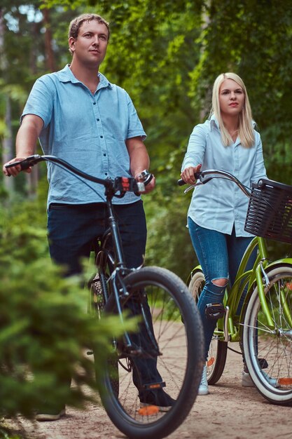 An attractive couple of a blonde female and man dressed in casual clothes on a bicycle ride in a park.