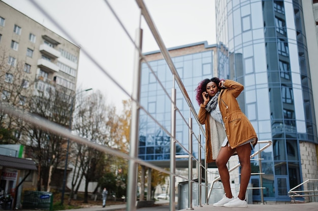 Attractive curly african american woman in brown coat posed near railings against modern multistory building speaking on mobile phone