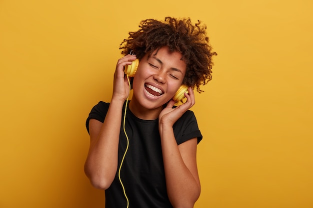 Attractive curly woman with toothy smiles, keeps hands on headset, dressed in black t shirt, isolated over yellow background