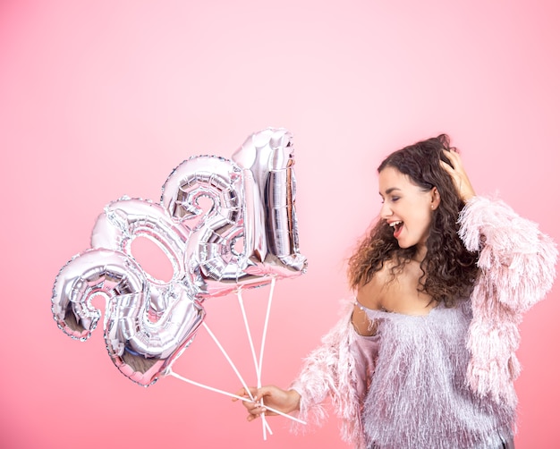 Attractive cute brunette girl with curly hair festively dressed posing on a pink background with silver balloons in hands for the new year concept