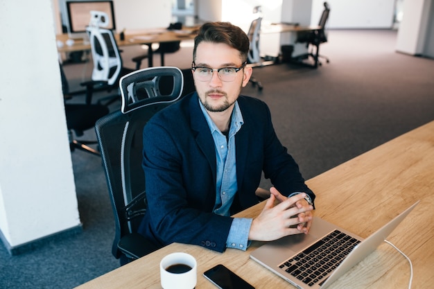 Free photo attractive  dark-haired man is sitting at the table in office. he wears blue shirt with black jacket. he is looking to the side. view from above.