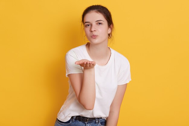 Attractive dark haired young woman dresses white t shirt blowing air kiss while posing