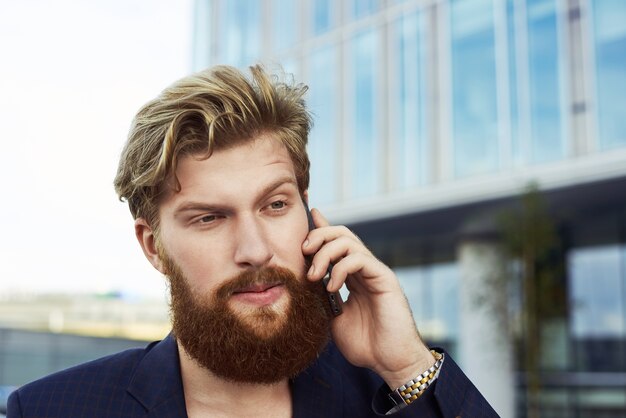 Attractive doubting man talk by mobile phone and walk outdoor near business buildings. Serious person in suit.