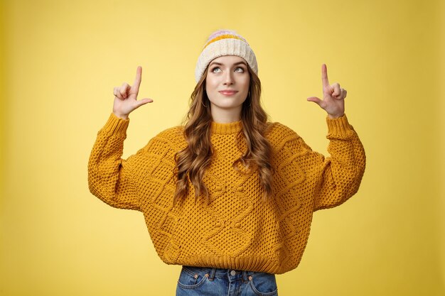 Attractive dreamy stylish college girl wearing hat warm sweater looking up pointing top smiling deli...