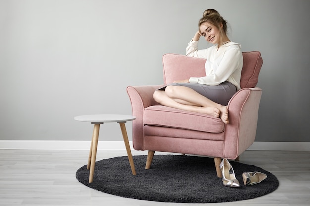 Attractive fashionable young European female with hair bun and bare feet sitting comfortably in pinkish armchair and smiling, enjoying spare time on her own, stylish high heeled shoes on carpet