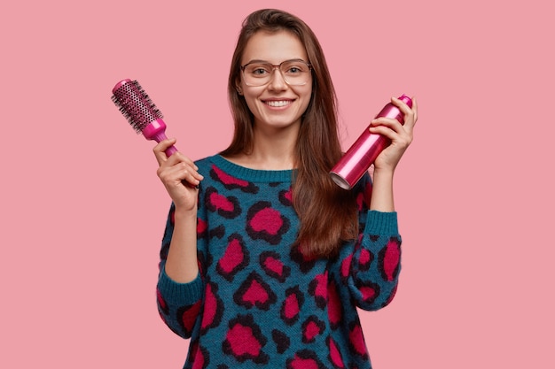 Free photo attractive female hairdresser holds comb and hairspay, wears big spectacles and sweater