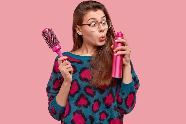 Free photo attractive female wears big square glasses, stares with stupefaction at hairspray, holds hairbrush, wants to make coiffure for special occasion