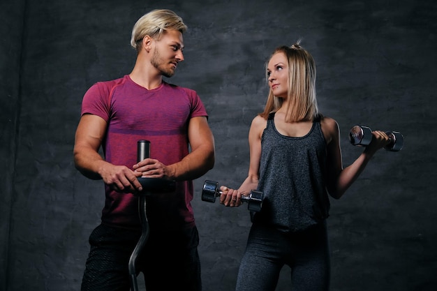 An attractive fitness couple of sporty male holds barbell and slim blond female holds dumbbells over grey background