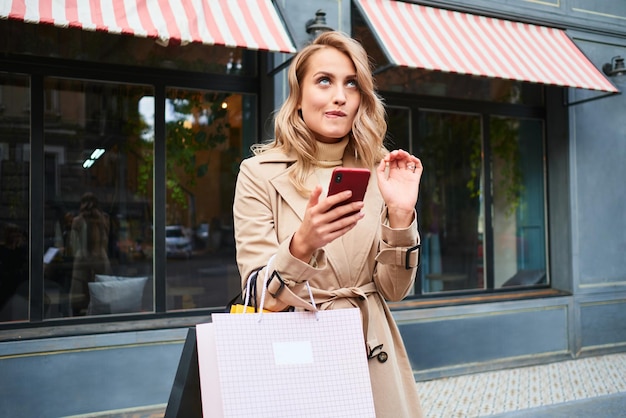 Attractive flirty blond girl in stylish trench coat with shopping bags dreamily using cellphone on city street