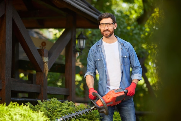 Free Photo attractive gardener posing while trimming conifer bush by electric hedge clippers in sunny day