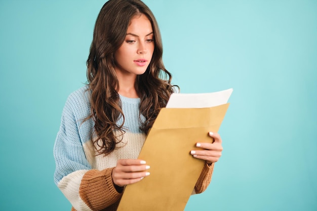 Attractive girl in cozy sweater thoughtfully opening response envelope over blue background