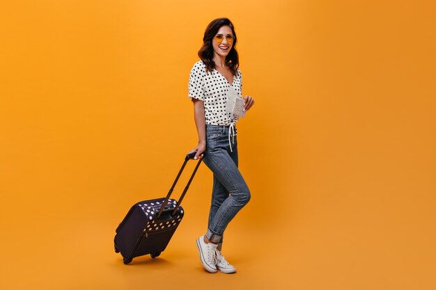 Attractive girl in glasses carries suitcase on orange background. Brunette with wavy hair in sunglasses in white blouse with black polka dots posing.