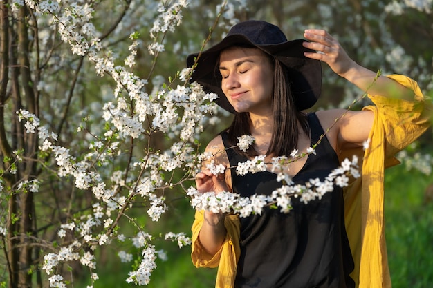 Free photo an attractive girl in a hat among blooming trees enjoys the smell of spring flowers