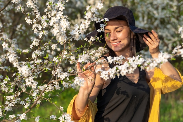 An attractive girl in a hat among blooming trees enjoys the smell of spring flowers