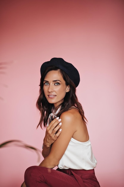 Attractive girl in hat and stylish outfit looking at camera with smile Pretty woman with beautiful eyes with white manicure posing on pink background