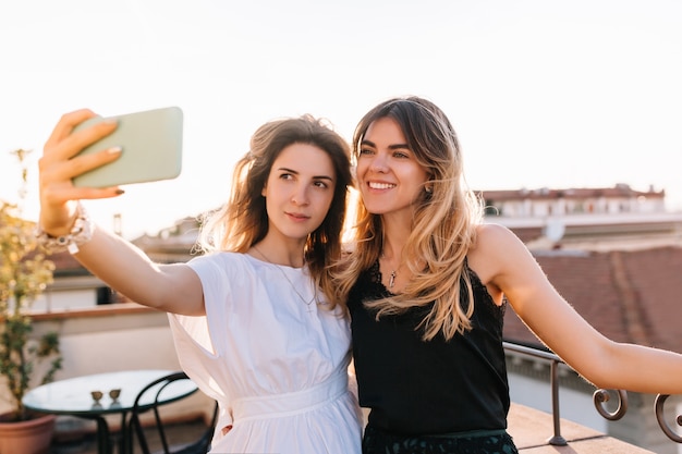 Attractive girl wearing white dress and trendy accessories taking selfie with best friend in outdoor cafe in morning