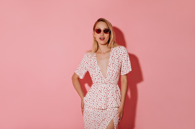 Free photo attractive girl with blonde hair in red sunglasses and earrings in stylish white dress looking into camera on isolated pink backdrop