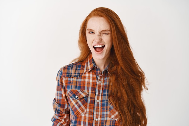 Free photo attractive girl with ginger hair and blue eyes winking at front assuringly, standing over white wall