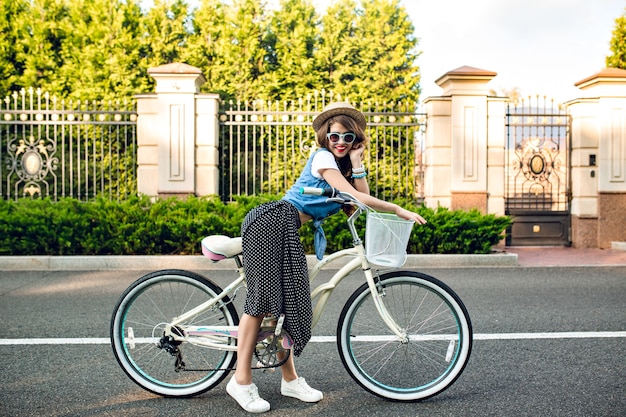 Free photo attractive girl with long curly hair in hat posing with bike on road. she wears long skirt, jerkin, blue sunglasses. she is smiling to camera.