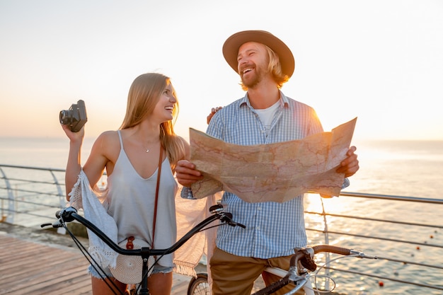 Free photo attractive happy couple traveling in summer on bicycles, man and woman with blond hair boho hipster style fashion having fun together, looking in map sightseeing taking photos on camera