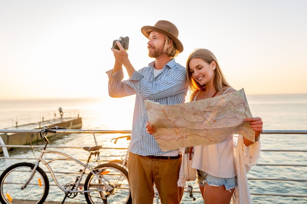 Free photo attractive happy couple traveling in summer on bicycles, man and woman with blond hair boho hipster style fashion having fun together, looking in map sightseeing taking photos on camera