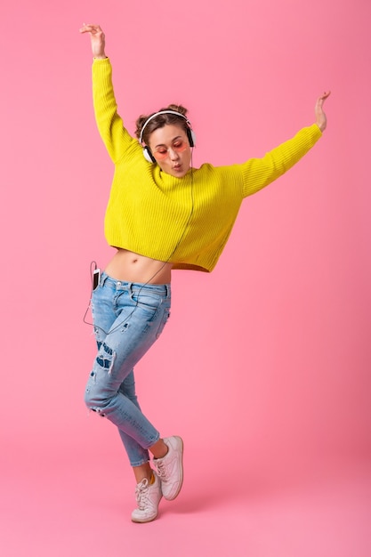 Free photo attractive happy funny woman dancing listening to music in headphones dressed in hipster colorful style outfit isolated on pink wall, wearing yellow sweater and sunglasses, having fun