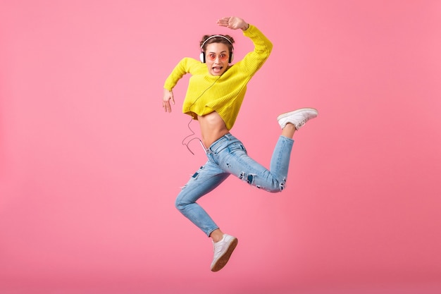 Free photo attractive happy funny woman jumping listening to music in headphones dressed in hipster colorful style outfit isolated on pink wall, wearing yellow sweater and sunglasses, having fun