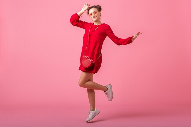 Attractive happy smiling stylish woman in red trendy dress jumping running on pink wall isolated, spring summer fashion trend, romantic mood flirty girl