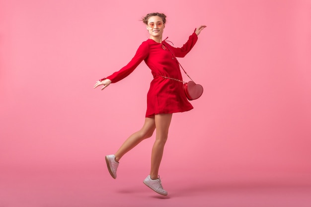 Attractive happy smiling stylish woman in red trendy dress jumping running on pink wall isolated, spring summer fashion trend, romantic mood flirty girl