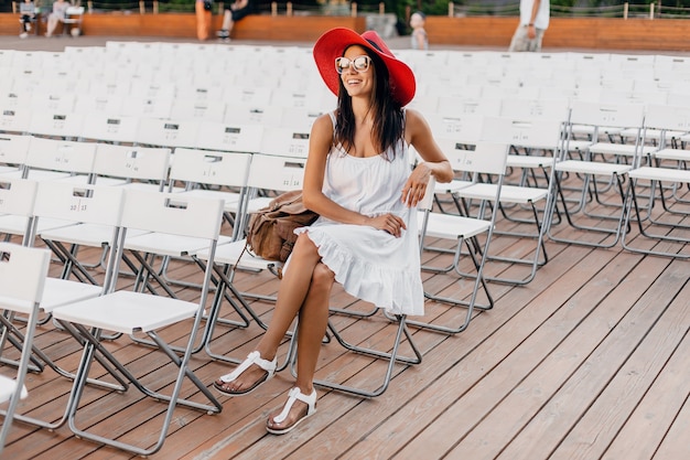 Attractive happy smiling woman dressed in white dress, red hat, sunglasses sitting in summer open air theatre alone, many chairs, spring street style fashion trend, waving hand hello