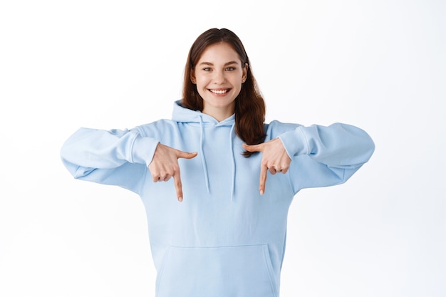 Free Photo attractive hipster girl with happy smile showing promotional text advertisement, pointing fingers down at logo, inviting check out promo, standing over white wall