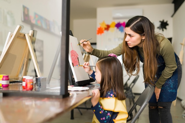 Attractive hispanic teacher is teaching a pretty child girl how to use the paint brush on a canvas during an art class