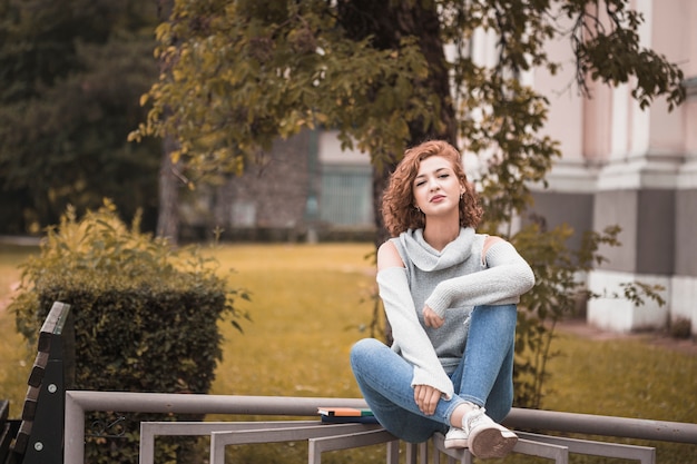 Free photo attractive lady in street wear sitting on rail and holding leg