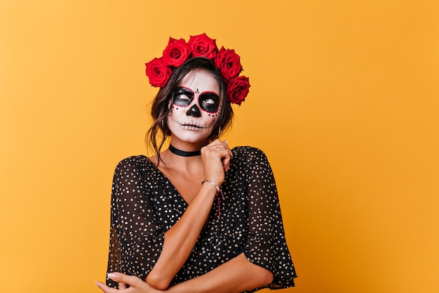 Attractive lady with Halloween makeup looks up thoughtfully. Portrait of girl with red flowers in her hair posing on orange background.