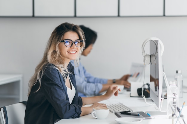 Free photo attractive laughing freelancer woman posing with cup of coffee at her workplace. chinese student in blue shirt works with document in campus with blonde friend in glasses.