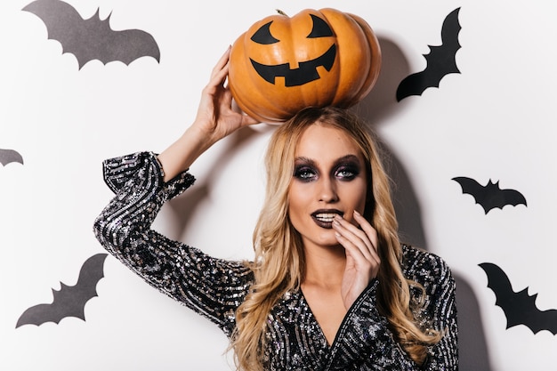 Attractive long-haired girl standing on white wall with bats. Indoor photo of appealing vampire with halloween pumpkin.