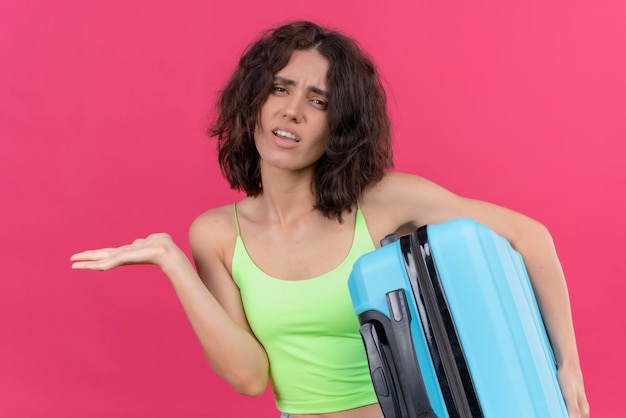 An attractive lovely woman with short hair wearing green crop top showing open hand palms holding blue suitcase