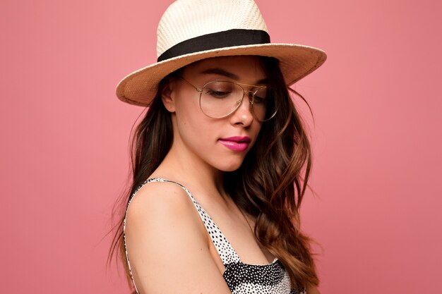 Attractive lovely woman with wavy hair in hat and glasses with pink lips posing over pink wall