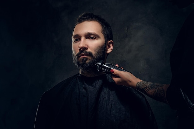 Free photo attractive man have beard trimming with machine at dark barbershop from female.