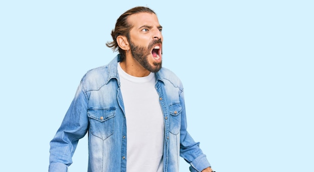 Attractive man with long hair and beard wearing casual denim jacket angry and mad screaming frustrated and furious, shouting with anger. rage and aggressive concept.