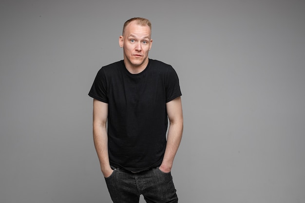 Attractive man with short fair hair wearing a black t-shirt and jeans keeping hands in pockets and surprising of something