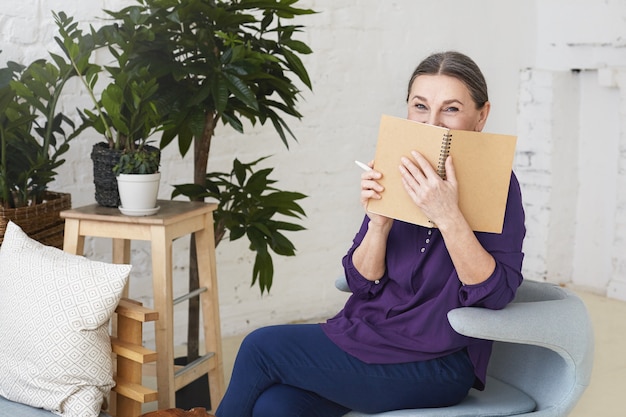 Attractive middle aged housewife on retirement siting on modern armchair in stylish living room interior, smiling and covering face with copybook while writing down grocery list before shopping