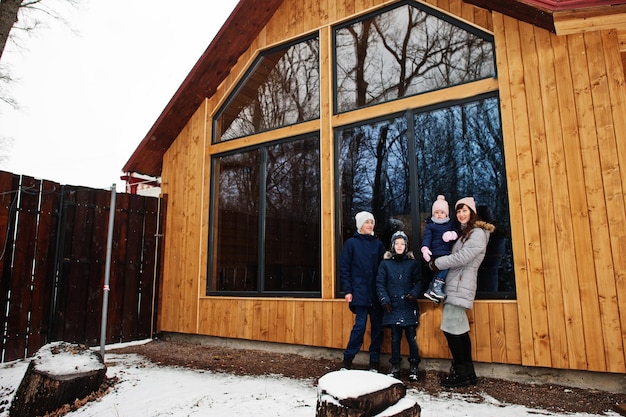 Attractive mother with her kids in winter day stand near wooden house with large window